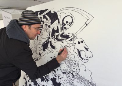 Cartoonists Simon Chadwick is wearing a beanie hat, standing up, and drawing a black and white cartoon of a skeleton riding a skeletal creature onto a white canvas