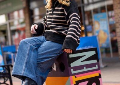 A woman sat on a colourful box, it says listen on the side. She is wearing headphones
