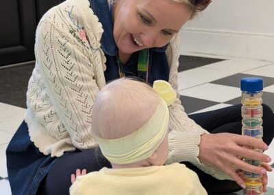 An adult sits on the floor interacting with a baby, holding a clear rattle toy filled with coloured pieces while the baby looks on.