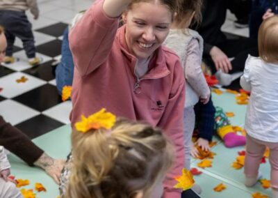 An adult sits on a play mat with several small children, holding up and dropping colourful leaves while children play around on a patterned floor.