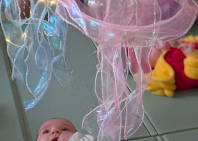A baby lies on a mat reaching up toward soft, translucent jellyfish-shaped decorations with lights hanging above.