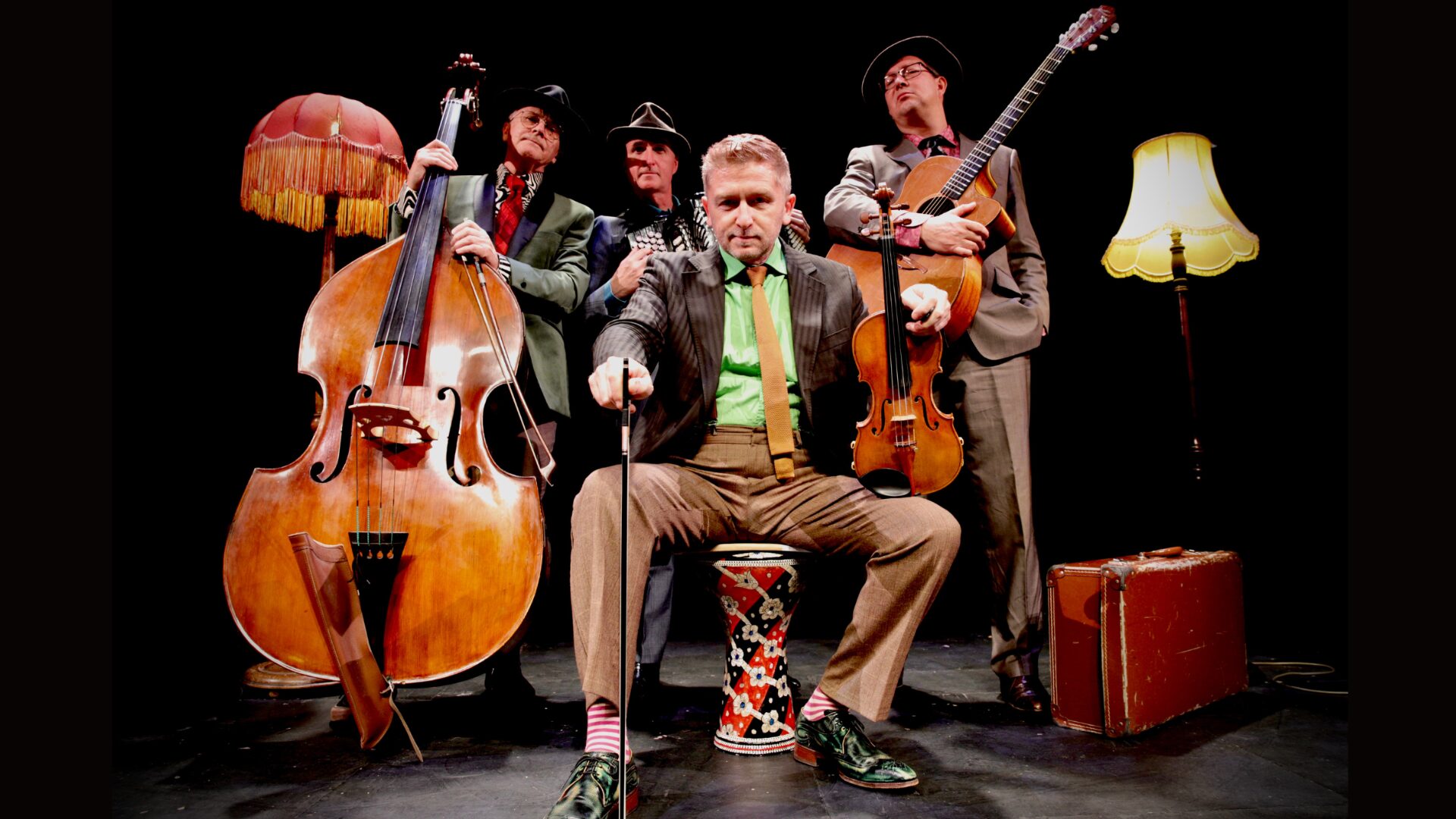 Group portrait of musicians with violin, accordion, guitar and double bass, seated and standing together against a dark backdrop in smart concert attire