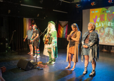Group of performers stood on stage looking out at the audience. There is a rainbow pride flag in the background