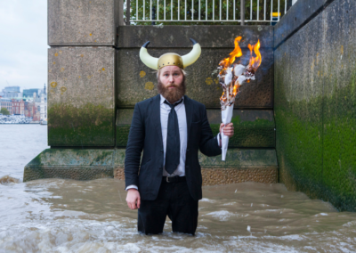 Performer in formal clothing stands on a dark stage holding a flaming torch aloft, creating a striking theatrical image that suggests storytelling, ritual and adventure.