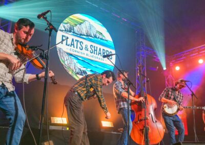 Five musicians perform on stage under colourful lights, playing fiddle, double bass, banjo and guitar before a large Flats & Sharps backdrop.