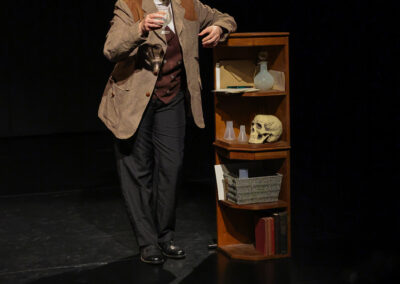 Solo performer beside a small cabinet of props, including books, glassware and a skull, during a theatrical storytelling performance.