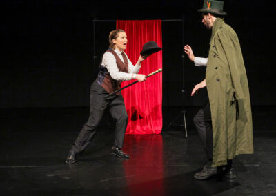 Performer brandishes a cane and hat opposite a cloaked character on a dark stage, with a red curtain creating a dramatic theatrical backdrop.